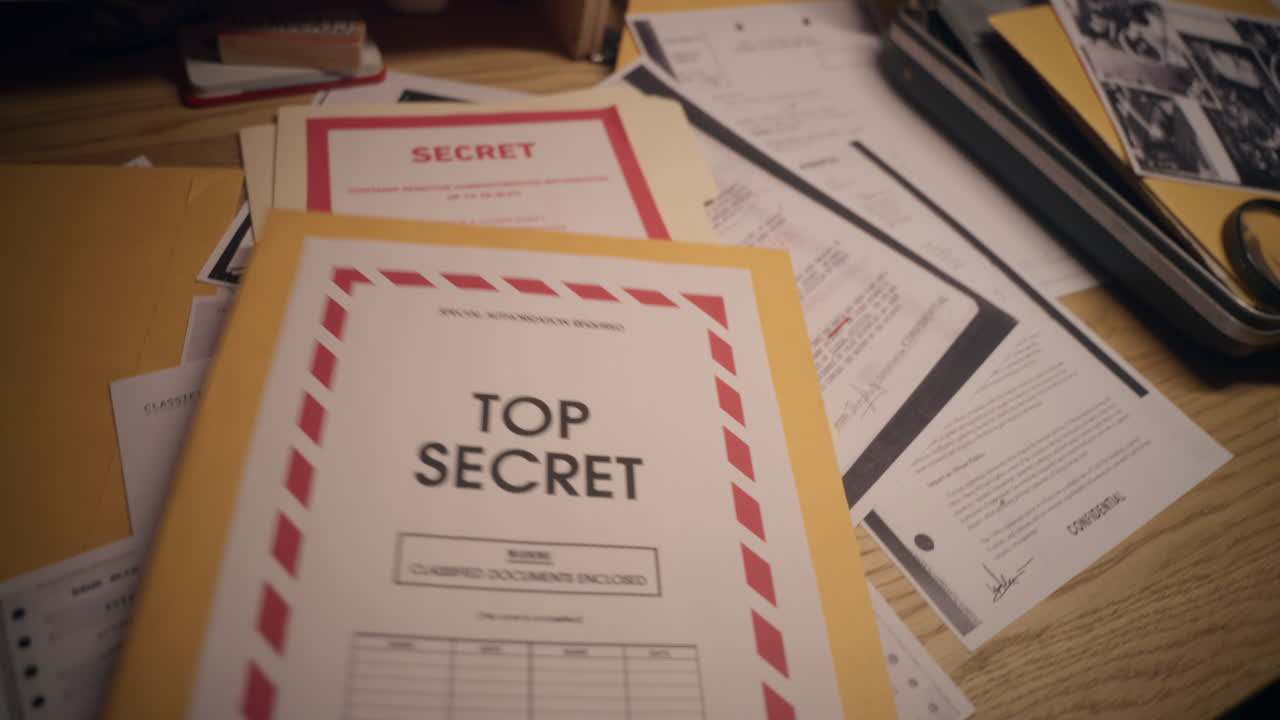 Cluttered desk covered in classified and top secret documents, including maps, photos, and redacted files. A briefcase and magnifying glass hint at espionage or intelligence work. Close-up