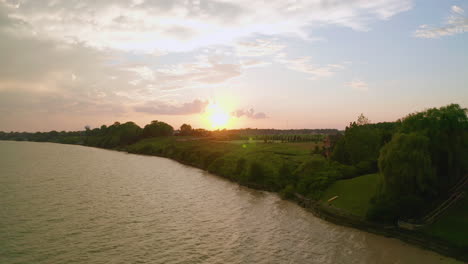 Eine-Drohne-Erhebt-Sich-Aus-Dem-Wasser-Und-Gibt-Den-Blick-Auf-Felder,-Vororte-Und-Hütten-Frei,-Während-In-Der-Ferne-Die-Sonne-Untergeht