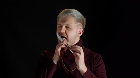 Handsome bearded man in stylish blouse licking knife on black background, Halloween horror theme
