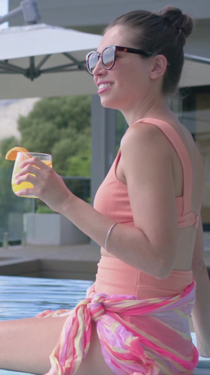 Donna a bordo piscina che allunga la mano per prendere una bevanda gialla, sorseggiando, rinfrescandosi e sorridendo, con icone di viaggio intorno al bicchiere.
