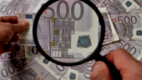 Person-checking-five-hundred-euro-banknote-for-authenticity-with-magnifying-glass,close-up