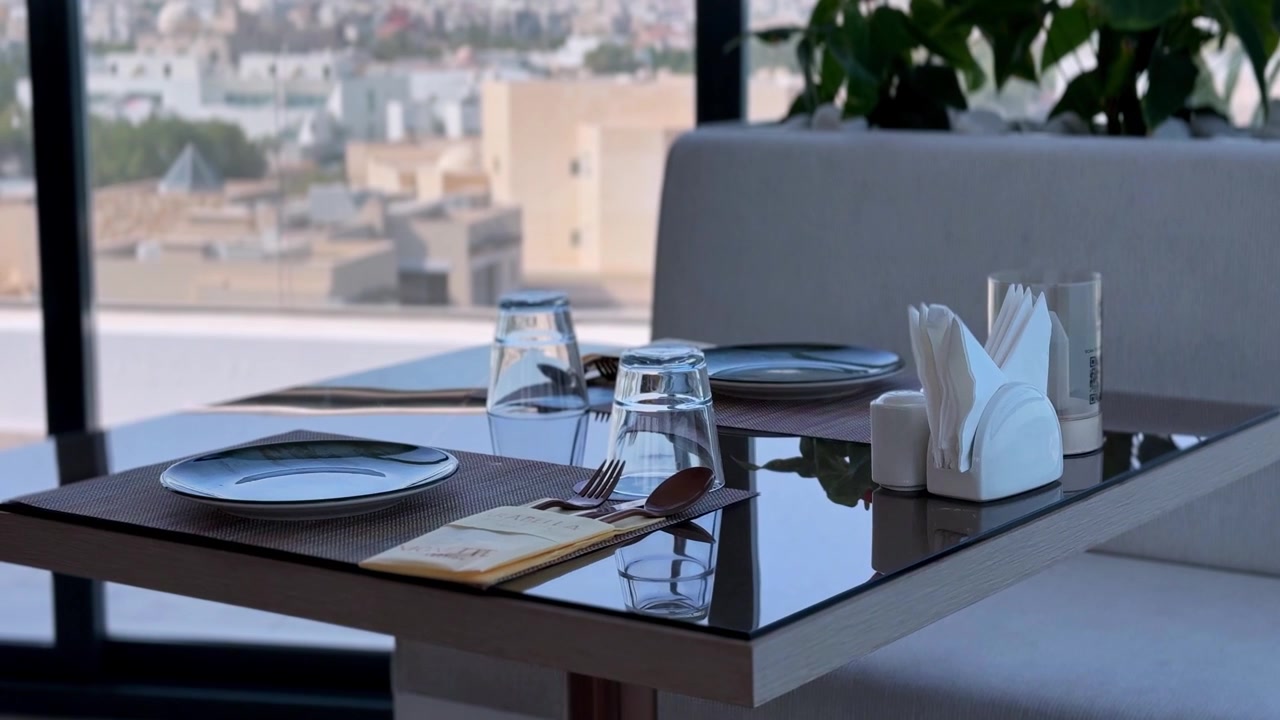 Close-up of an empty modern restaurant table setting prepared for daytime dining. The glossy black table features plates, cutlery, and glasses beside a large window overlooking a blurred city skyline.