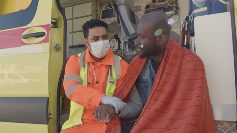 male paramedic taking the pulse and talking with an injured african-american patient while both sitting in the back of the ambulance.