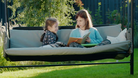 girl reading a book to her younger sister, relaxing on a swing in the backyard of the house