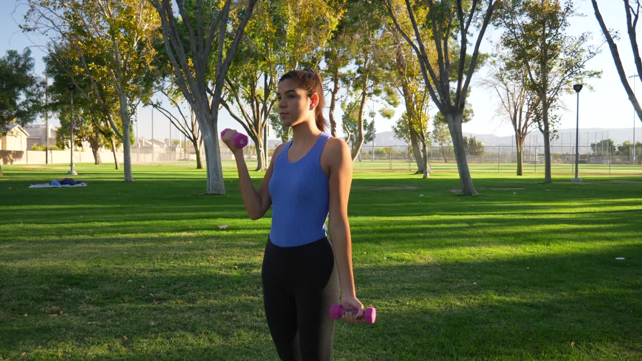una mujer joven haciendo un entrenamiento con mancuernas en el parque realizando rizos de bíceps para construir músculo del brazo y fuerza a cámara lenta