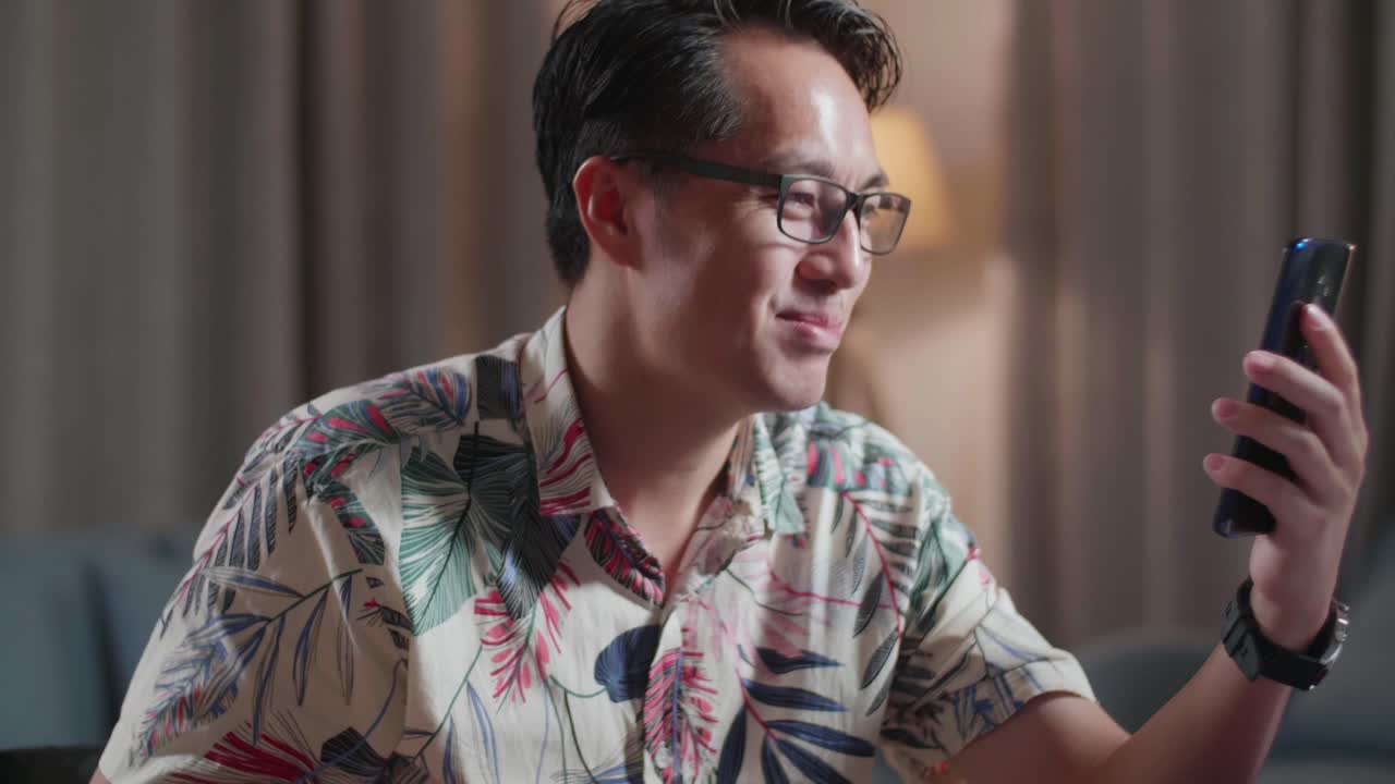 체어에 앉은 아시아인 남자가 손을 흔들며 스마트폰으로 비디오 통화를 하며 집에 있는 카메라 에 있는 데스크으로 비디오를 편집하고 있다.