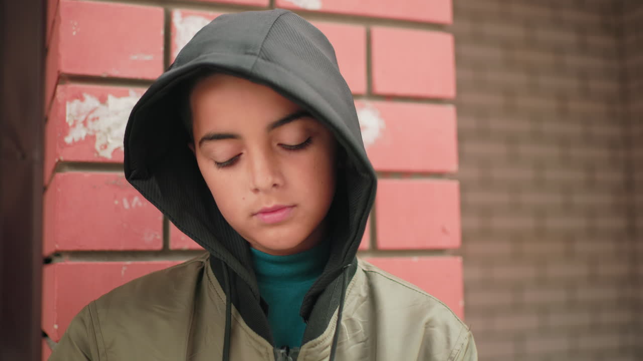 Close up of boy in hooded jacket standing against red brick wall lifting head up calmly then bringing it down again with neutral expression, outdoors near urban building background
