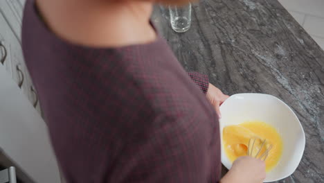 close-up of woman with golden hair wearing plaid gown stirring egg in bowl, she briefly lifts whisk before continuing to mix, background features a marble countertop