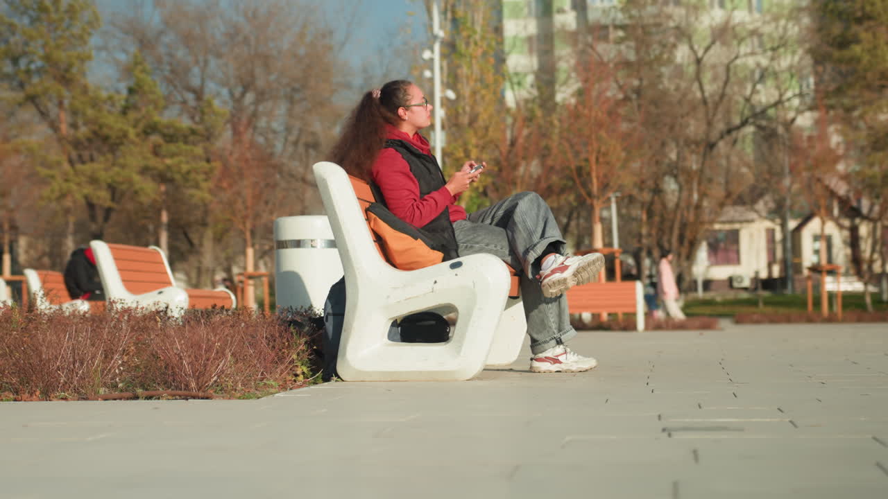 晴れた都会の公園のベンチでスマートフォンを操作する若い女性。カジュアルな服装にバックパックを背負い、通勤途中の短い休憩中。ヘッドホンが見え、遠くには歩行者がいる、リラックスした昼間の風景。