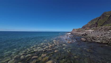 rugged rocky shoreline on coast of madeira island. clear blue water of the atlantic and steep cliffs seen in aerial view from drone flying low over gentle waves breaking on beach.