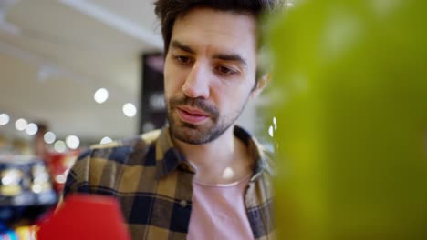 Close-up-a-confident-brunette-guy-with-stubble-in-a-checkered-shirt-approaches-the-counter-in-a-supermarket-and-chooses-the-red-product-he-needs