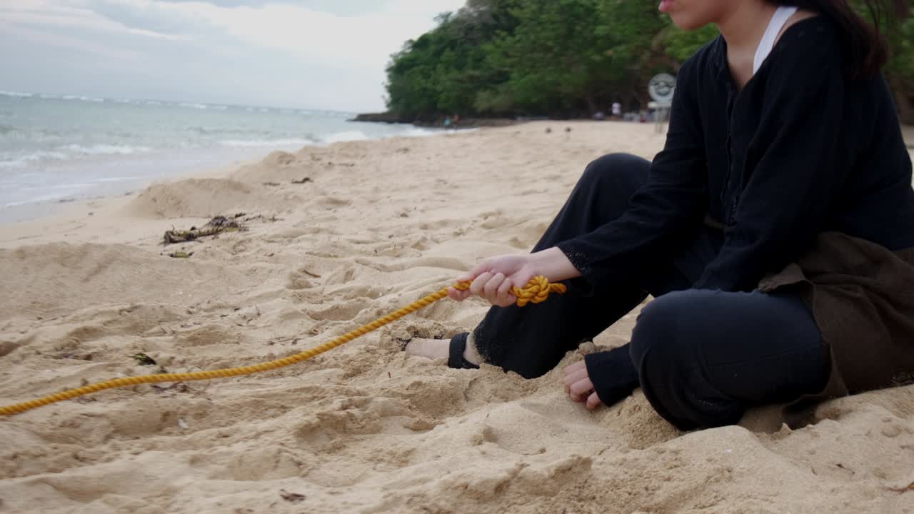 Static shot of female teenager sitting down by sandy beach grabbing rope leading to coastal ocean waters, representing depression and anxiety.
