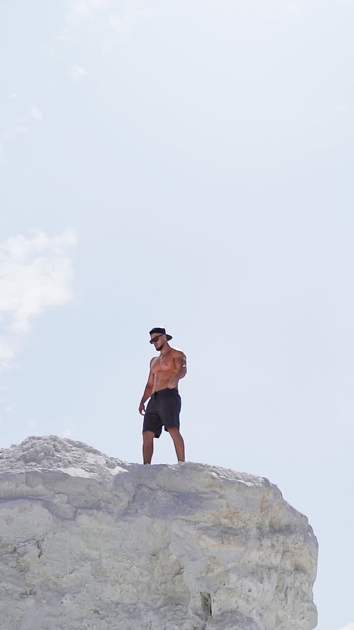 Sportive man on the edge of the hill. Shirtless man in shorts under the light sky. Muscular athlete on the top of mount in summer. View from below. Slow motion. Vertical video