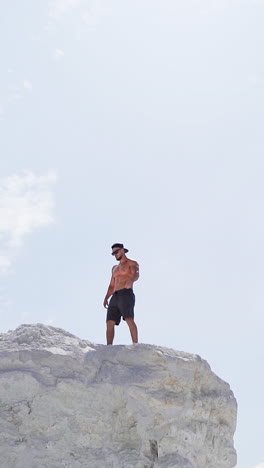 sportive man on the edge of the hill. shirtless man in shorts under the light sky. muscular athlete on the top of mount in summer. view from below. slow motion. vertical video