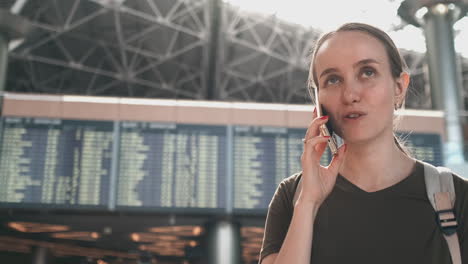 Parada-En-La-Pantalla-Con-Información-Sobre-La-Salida-Del-Avión-En-El-Aeropuerto,-Una-Joven-Con-Una-Mochila-Hablando-Por-Un-Teléfono-Móvil