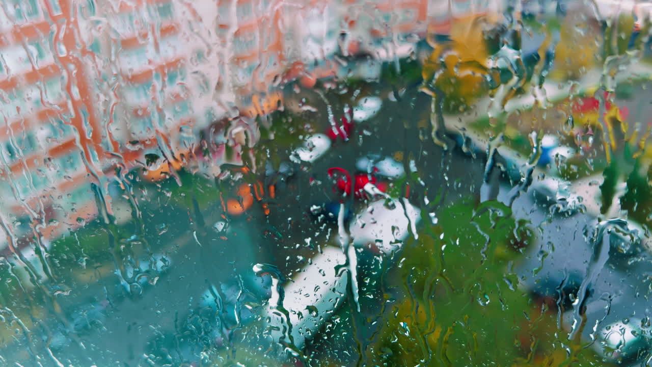 Looking at rainy day from home. Rain water dripping by the glass. Blurred houses and cars at backdrop.