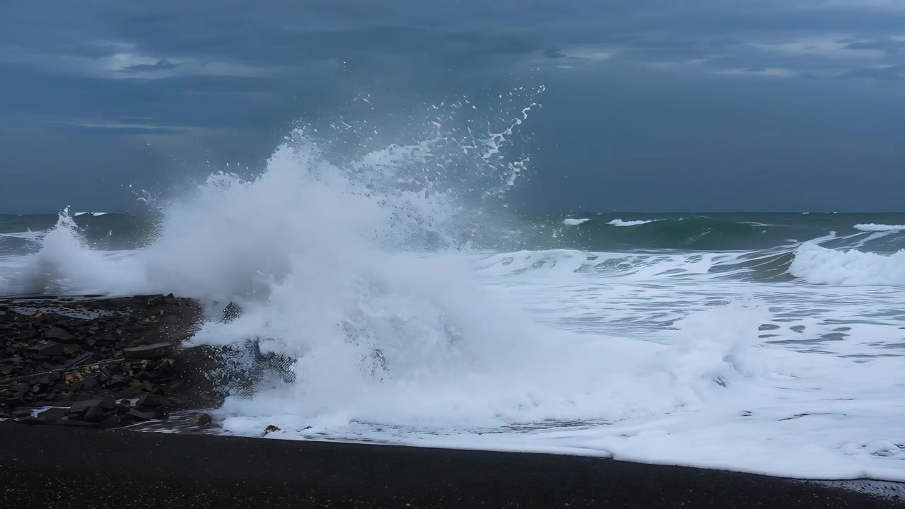 Crashing storm swell striking rocky jetty sending ocean wave spraying foam across dark sand shore