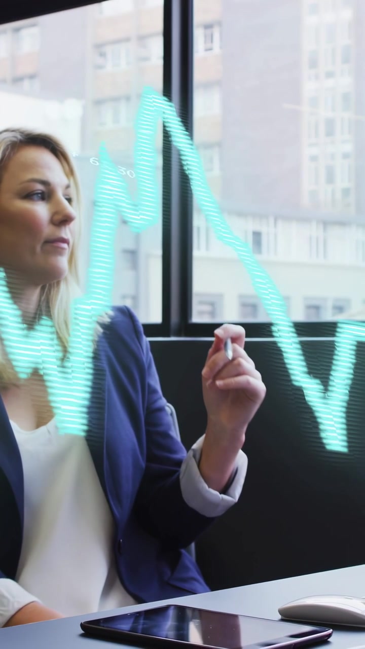 Video verticale: Donna alla scrivania vicino alla finestra, che usa il mouse, prende una penna e rivede una sovrapposizione di grafici finanziari.