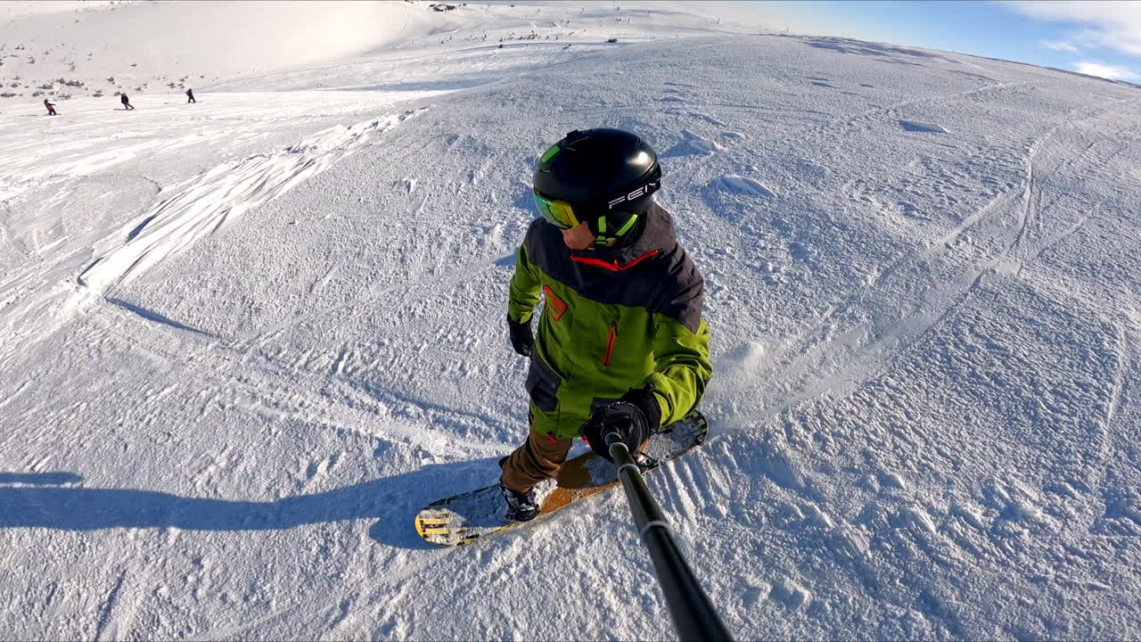 Sportsman going down the mountain slope taking selfie. Other people stand or ride at backdrop.