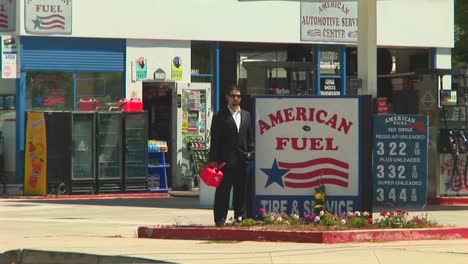 a man holding a gas can stands next to an american fuel gas station with traffic zipping by