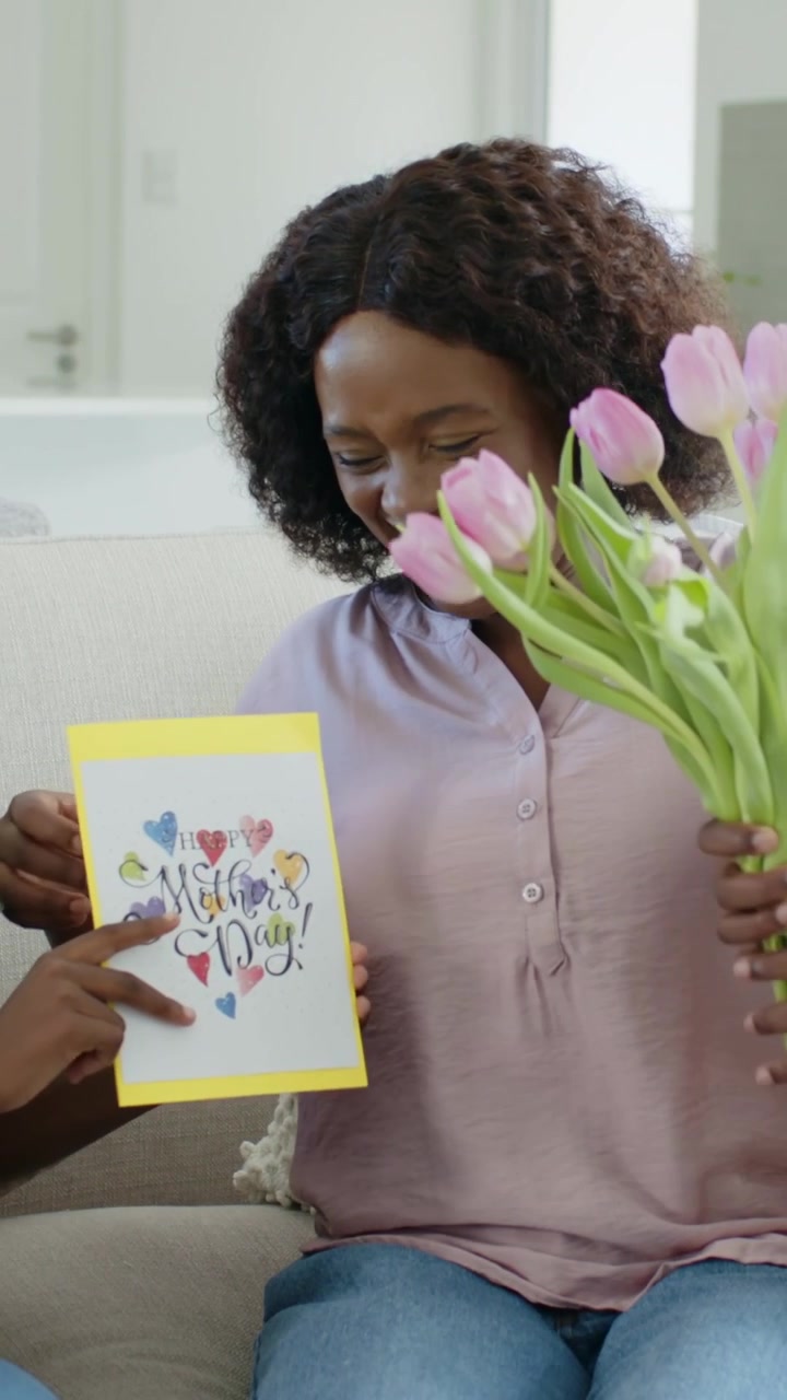 Video verticale: Mamma che ride con un top viola, legge un biglietto giallo mentre un bambino le porge dei tulipani rosa sul divano.