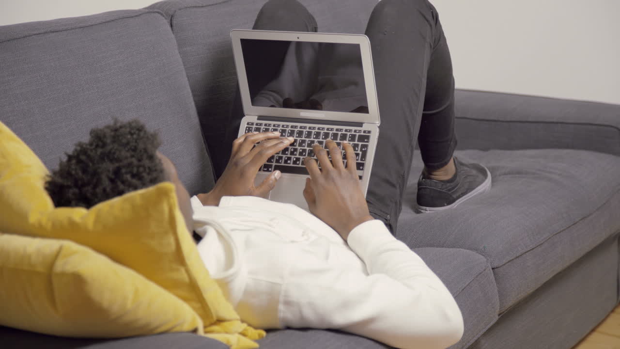 African American young man lying on couch and using laptop
