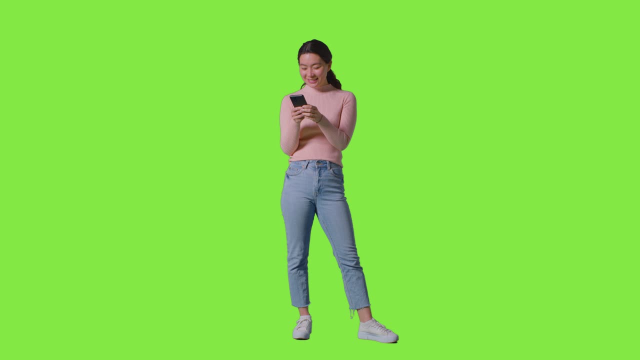 Ripresa in studio a figura intera di donna che sorride e ride a un messaggio o contenuto sul telefono cellulare contro uno schermo verde.