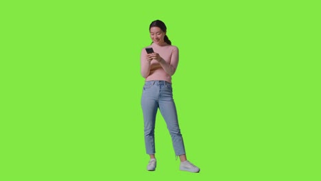 full length studio shot of woman smiling  and laughing at message or content on mobile phone against green screen