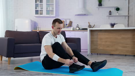 young man ties shoelace at home before the training. sportive guy sitting on a mat and dressing his black trainers for doing exercises.