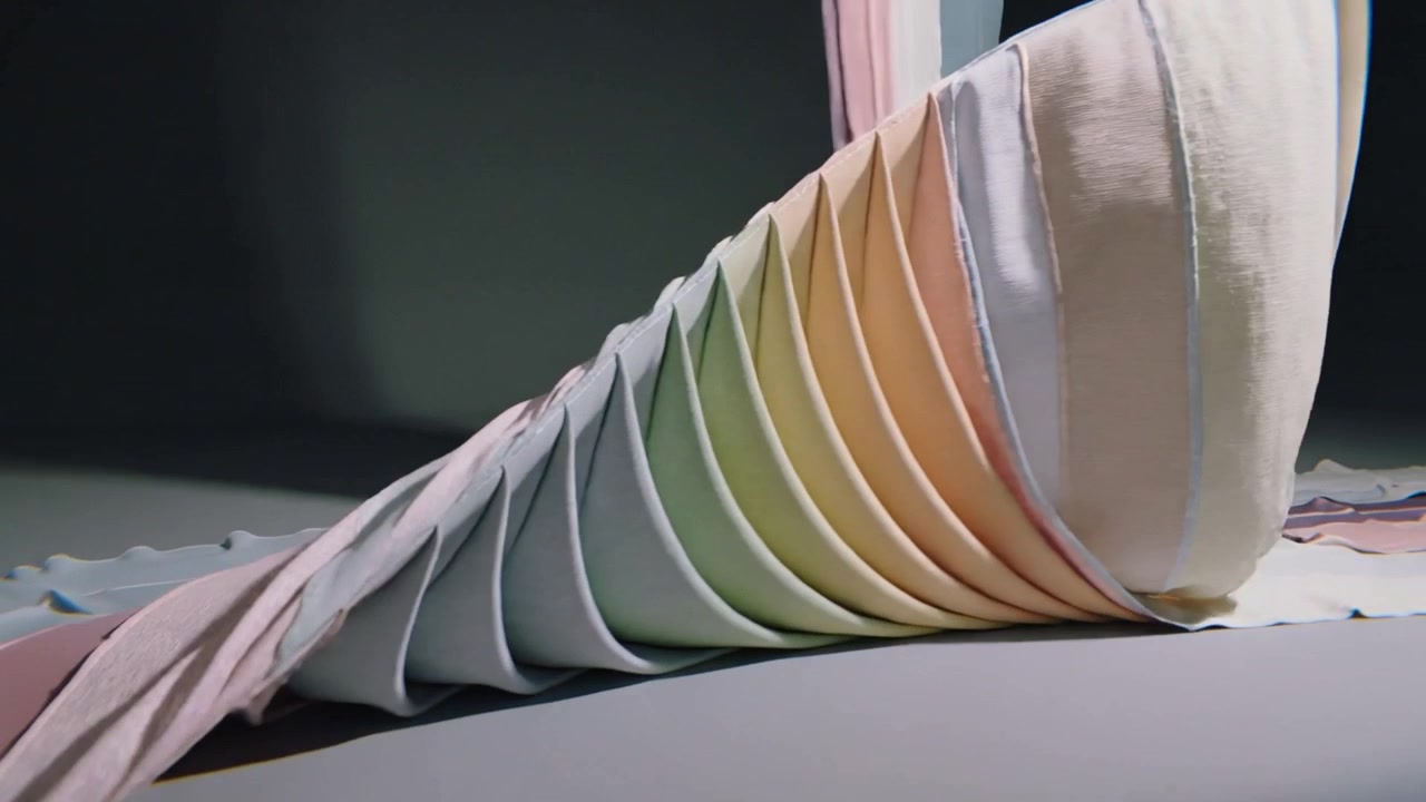 Tirando una striscia plissettata pastello, un'attrezzatura da studio forma un cono affusolato che viene poi drappeggiato su un cuneo per l'esposizione.