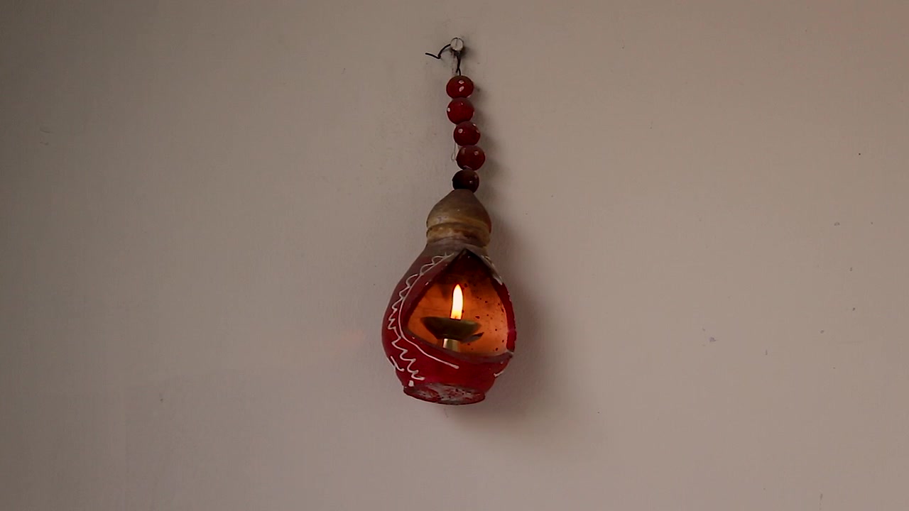Tradizionale lampada a olio in argilla sospesa con fiamma luminosa contro una parete semplice. Ideale per Diwali, spiritualità, meditazione, cultura indiana e progetti per un'atmosfera domestica pacifica