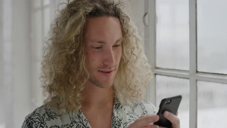 retrato de un atractivo joven estudiante usando un teléfono inteligente enviando mensajes de texto navegando en línea en las redes sociales leyendo mensajes en la tecnología de teléfonos móviles en un apartamento de pie junto a la ventana