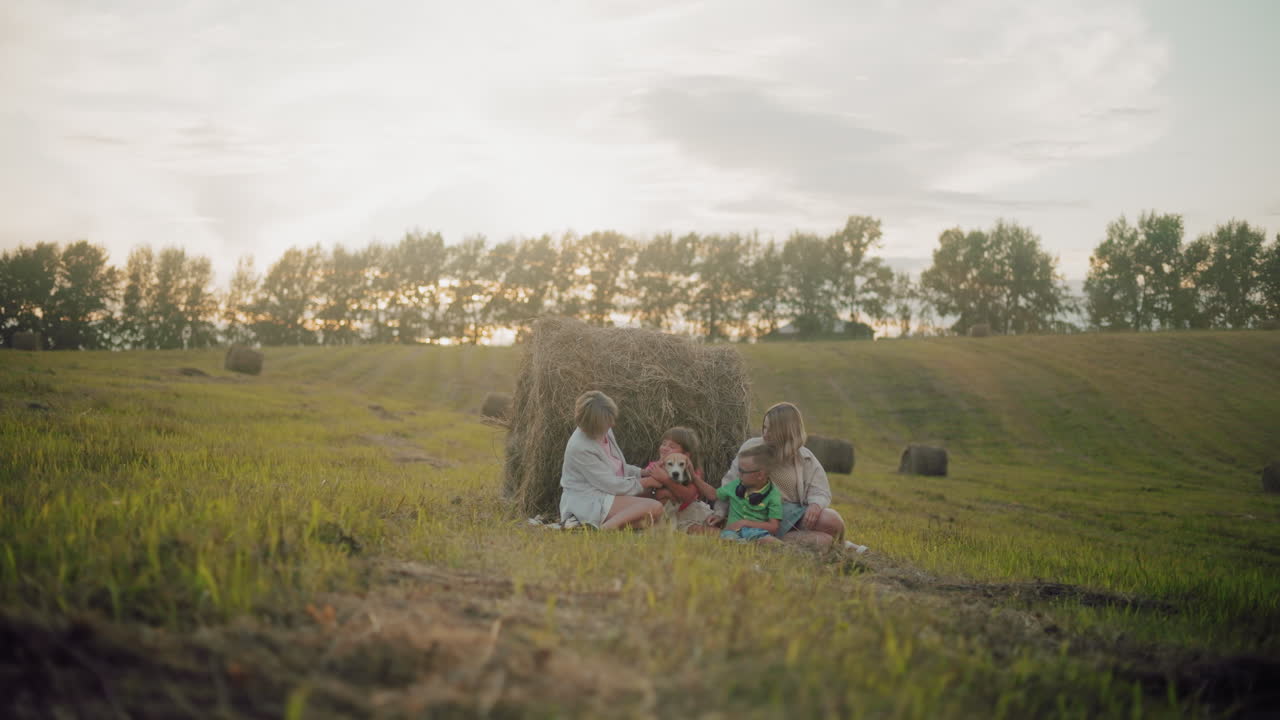 사랑하는 가족이 풀 근처의 야외에서 모여, 황금빛 해가 지는 아래 개를 만지고, 따뜻하고 즐거운 분위기는 강한 유대감, 행복과 평화로운 시골 풍경을 강조합니다.