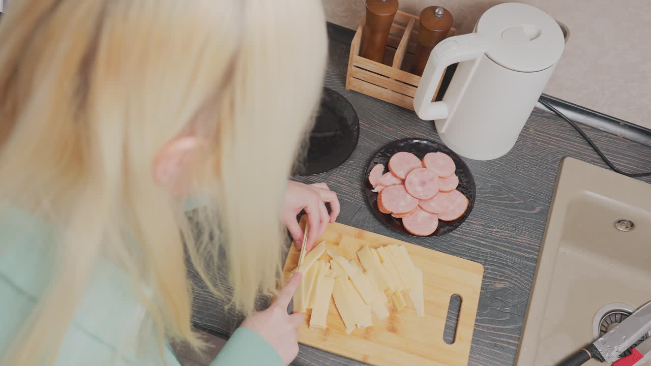 overhead view of blonde woman wearing green shirt slicing yellow cheese on wooden cutting board beside plate with sliced sausage, electric kettle, spice jars in wooden holder