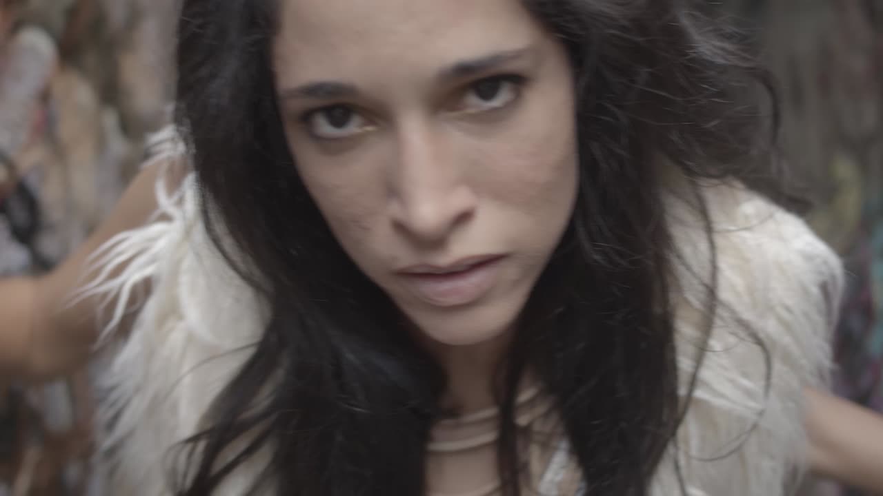 primer plano del rostro de una mujer con cabello largo, uñas negras y anillos de latón haciendo una danza interpretativa en un entorno urbano arenoso