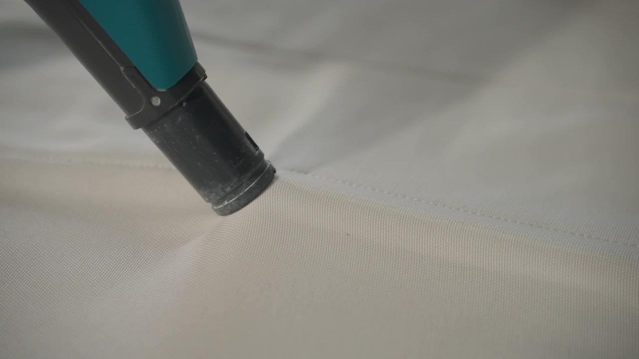 Vacuum nozzle cleaning mattress fabric closeup, detailed sequence showing suction head gliding over mattress seam lifting dust and light stains, soft neutral lighting, subtle motion, domestic house