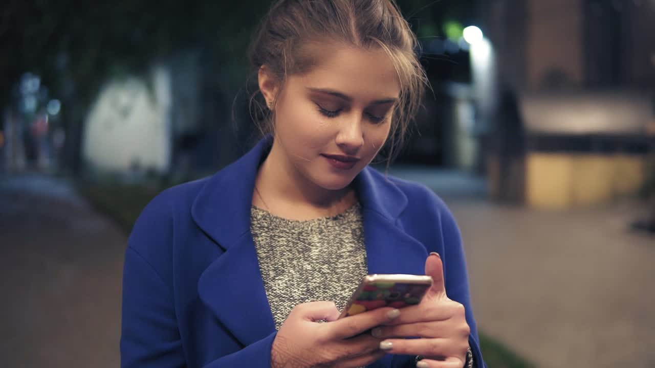 Young attractive woman using smartphone at night in city. Beautiful girl texting on smartphone outdoors. Tourist Girl with