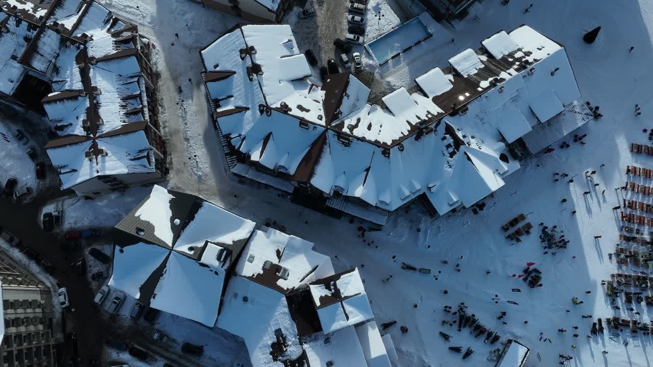 aerial shot of a small town square fully covered in snow. People are gathered in the streets enjoying various winter activities, while the surrounding buildings are topped with layers of snow