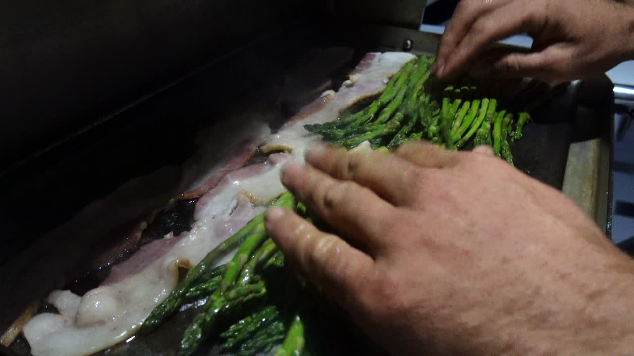 Sailboat captain cooks asparagus and bacon on a grill on the boat at night.