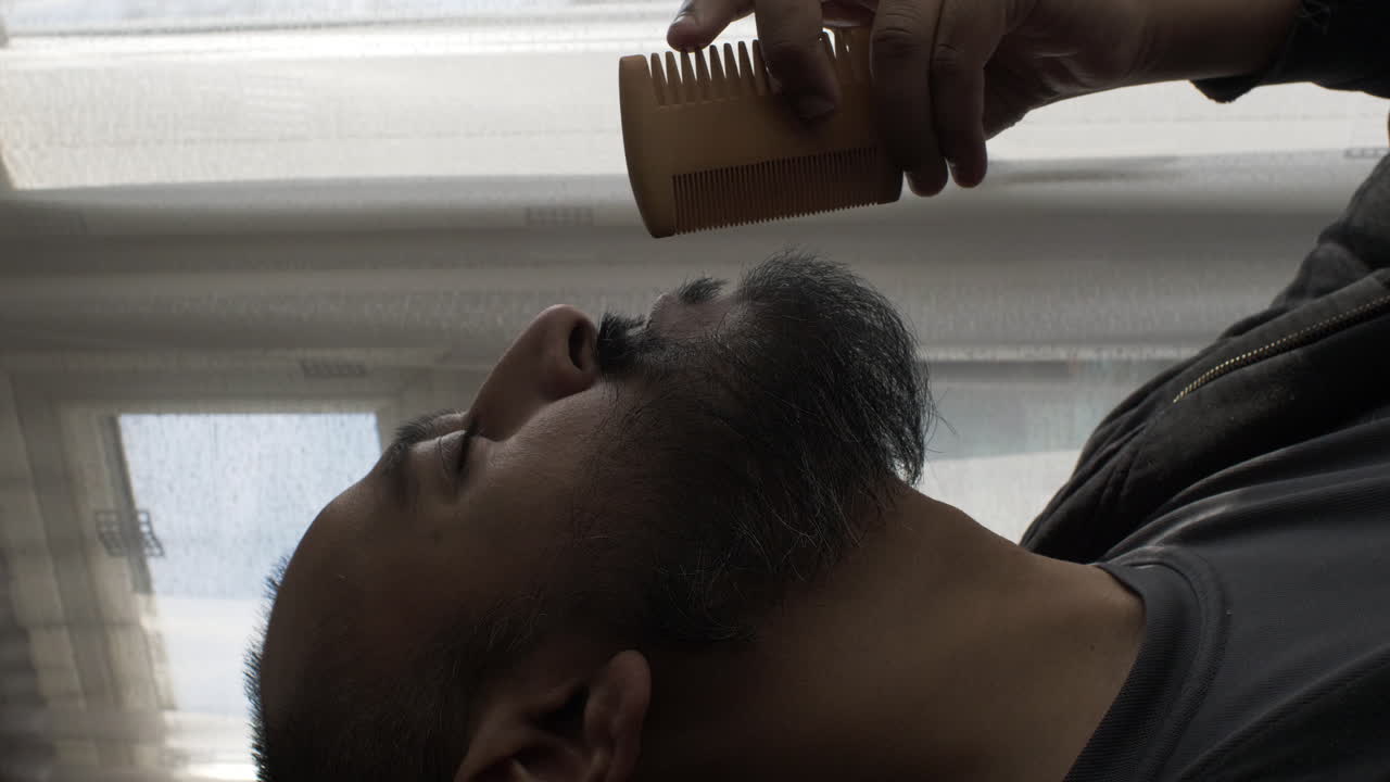 커튼 창문 근처에서 수염을 빗고 있는 소수 민족 성인 남성