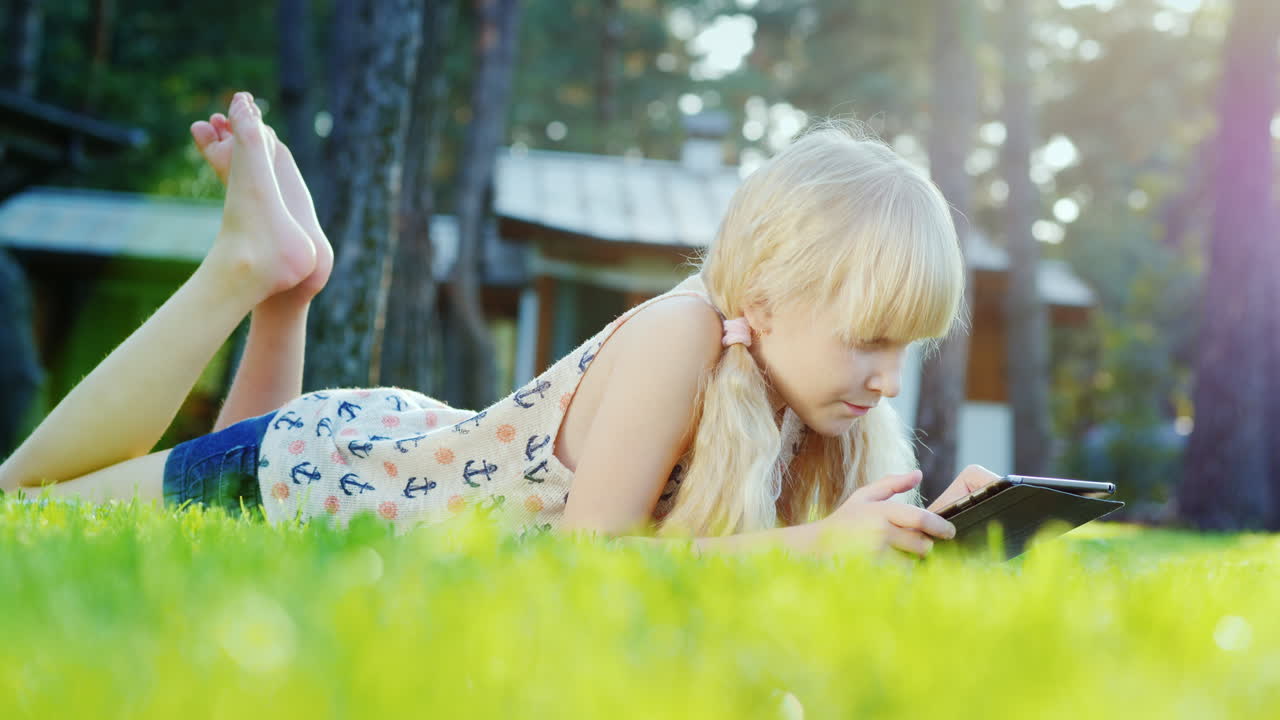 белокурая девочка 6 лет, играющая на планшете, эмоционально реагирует, лежа на газоне на заднем дворе дома.