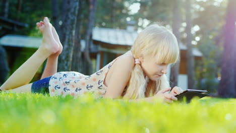 blonde girl 6 years playing on the tablet emotionally reacts lying on the lawn in the backyard of th