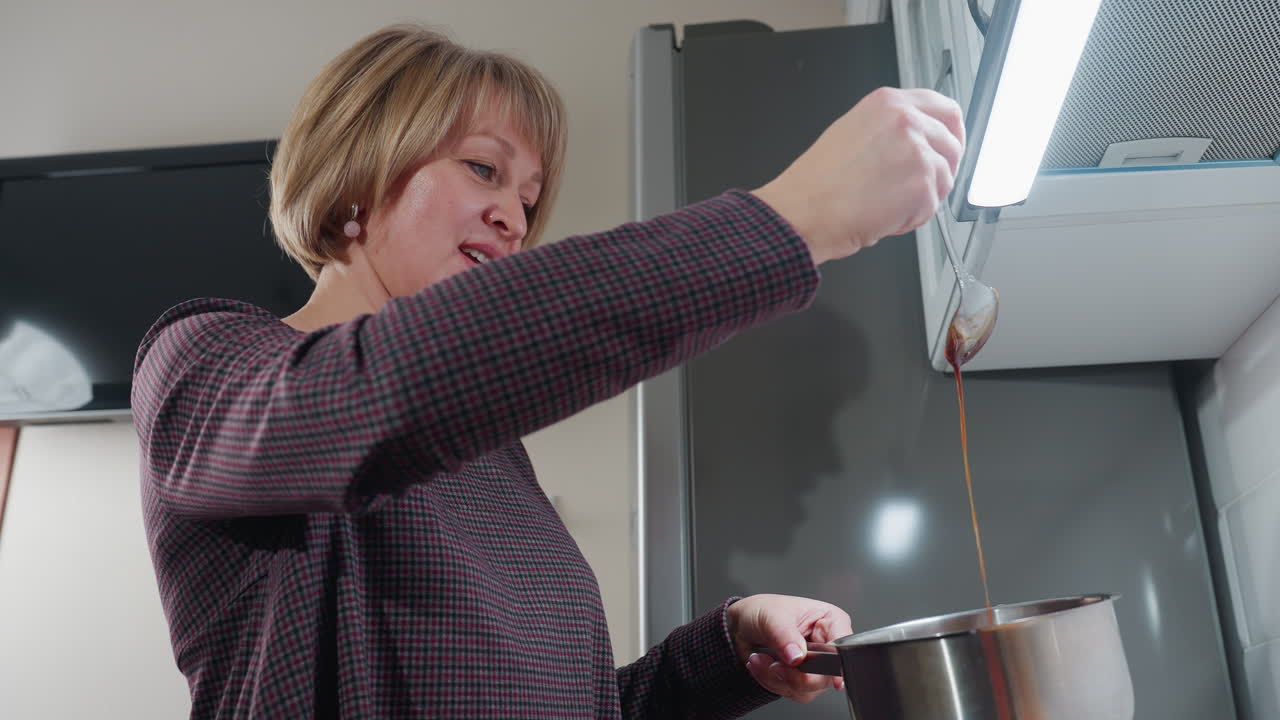 mujer rusa en ropa a cuadros graciosamente vierte sopa de la cuchara en la olla mientras baila en su cocina, la iluminación suave mejora el ambiente cálido con el reflejo de sombras en la pared