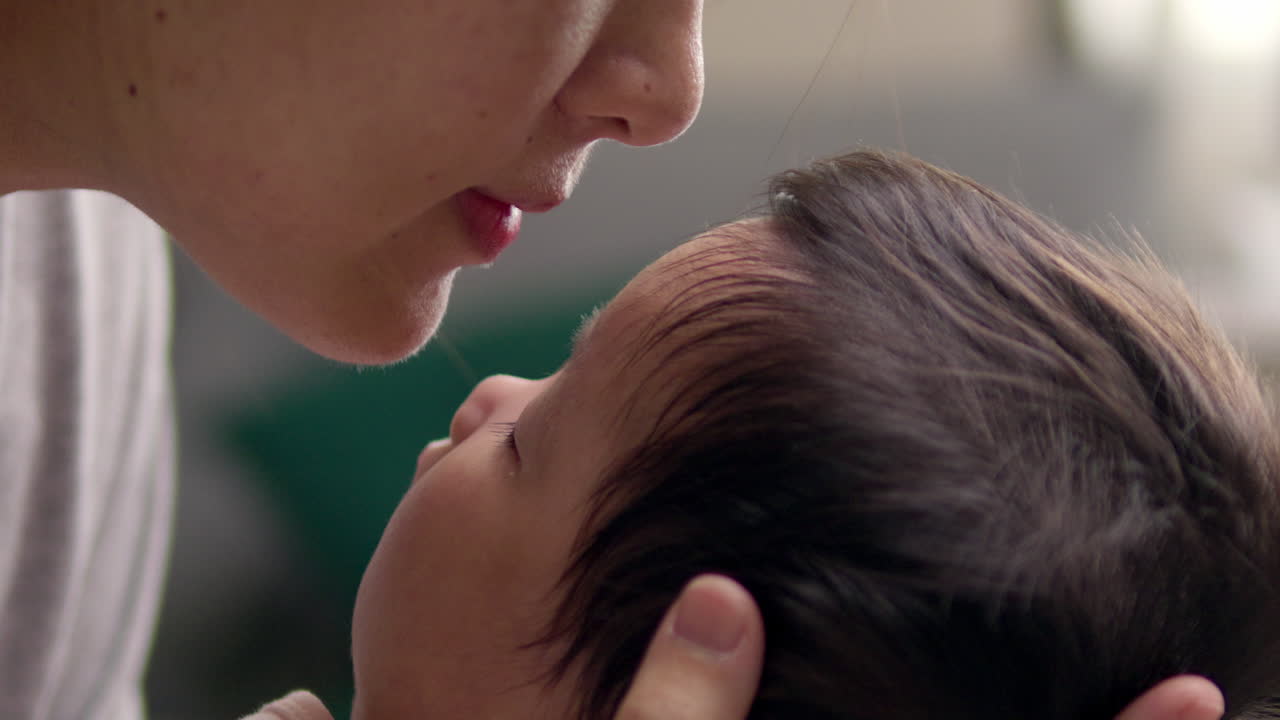 Panning from the right to the left side of the frame shows a newborn child's head cradled in her mother's hands, as her mother leans forward to kiss her gently.