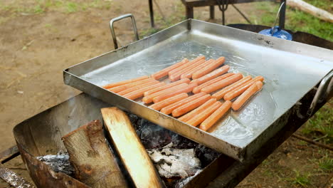 pouring olive oil over sausages in pan on open fire - campsite breakfast