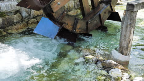 Close-up-of-wooden-water-mill-in-Slovenia,-lake-Jasna,-beautiful-colorful-stream-pushing-circle-around-with-lots-of-rocks-on-side