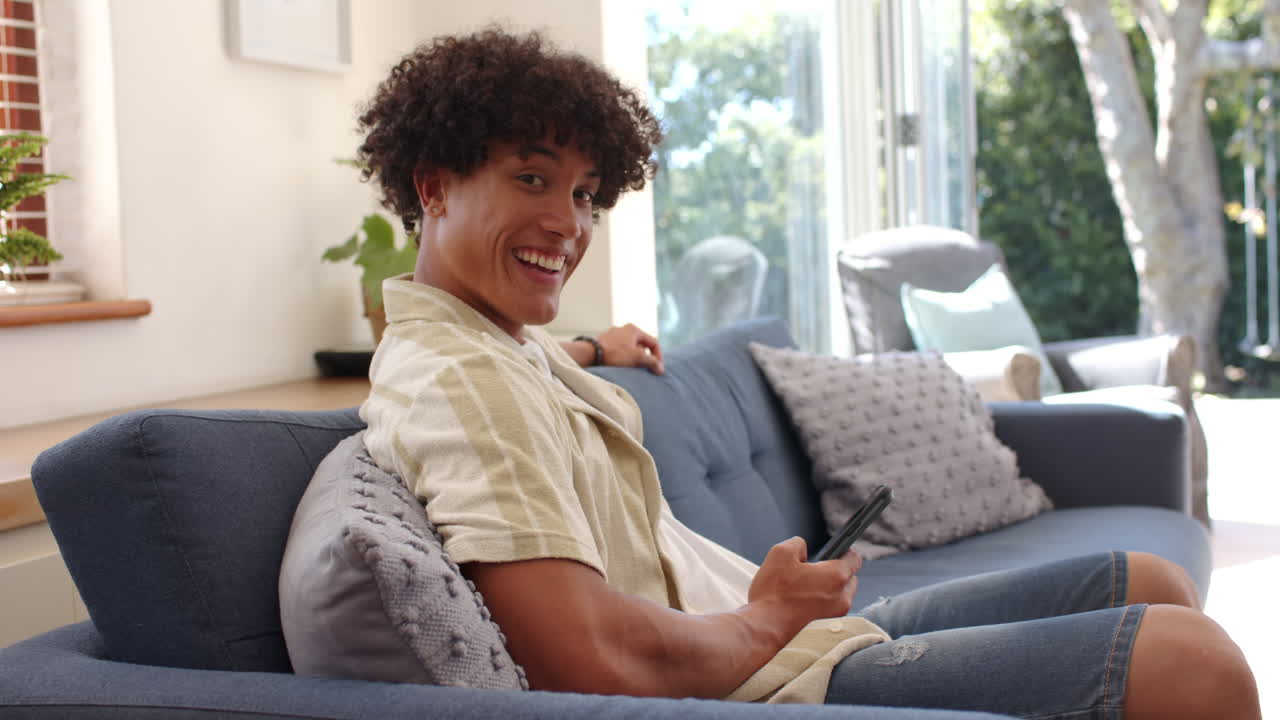 Sitting on couch, young man holding smartphone and smiling at camera, at home