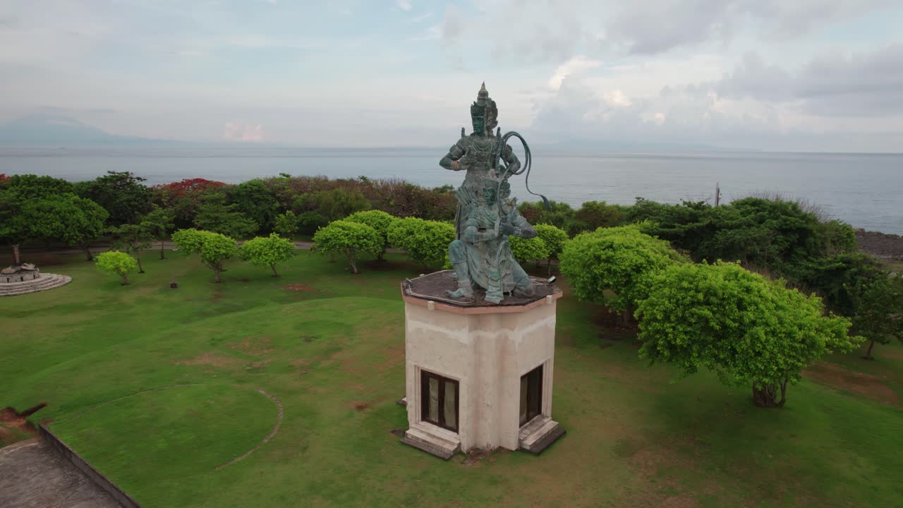 Drone video showing large mythological statue with bow standing on coastal pedestal in landscaped park near Garuda Wisnu Kencana Cultural Park in Bali, creating grand cultural scene