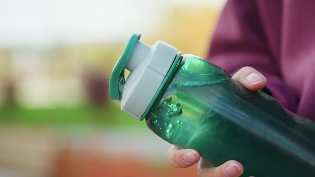 Female hand gripping translucent green water bottle with visible swirl and condensation motion against blurred outdoor location conveying dynamic hydration and fitness habit energy in urban scene