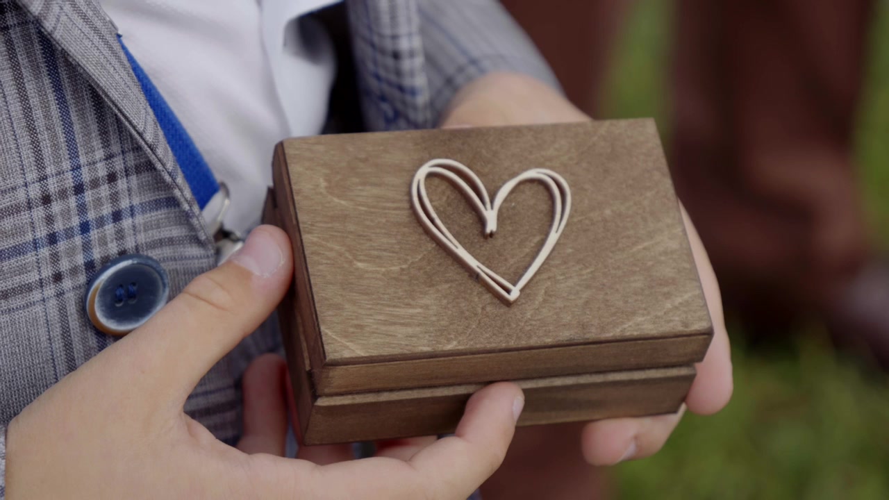 A young boy is holding a wooden box with a heart symbol, inside are wedding rings